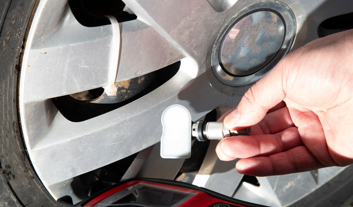 Technician holding a white TPMS sensor with metal valve stem next to an alloy car wheel, with a TPMS programming tool in the foreground