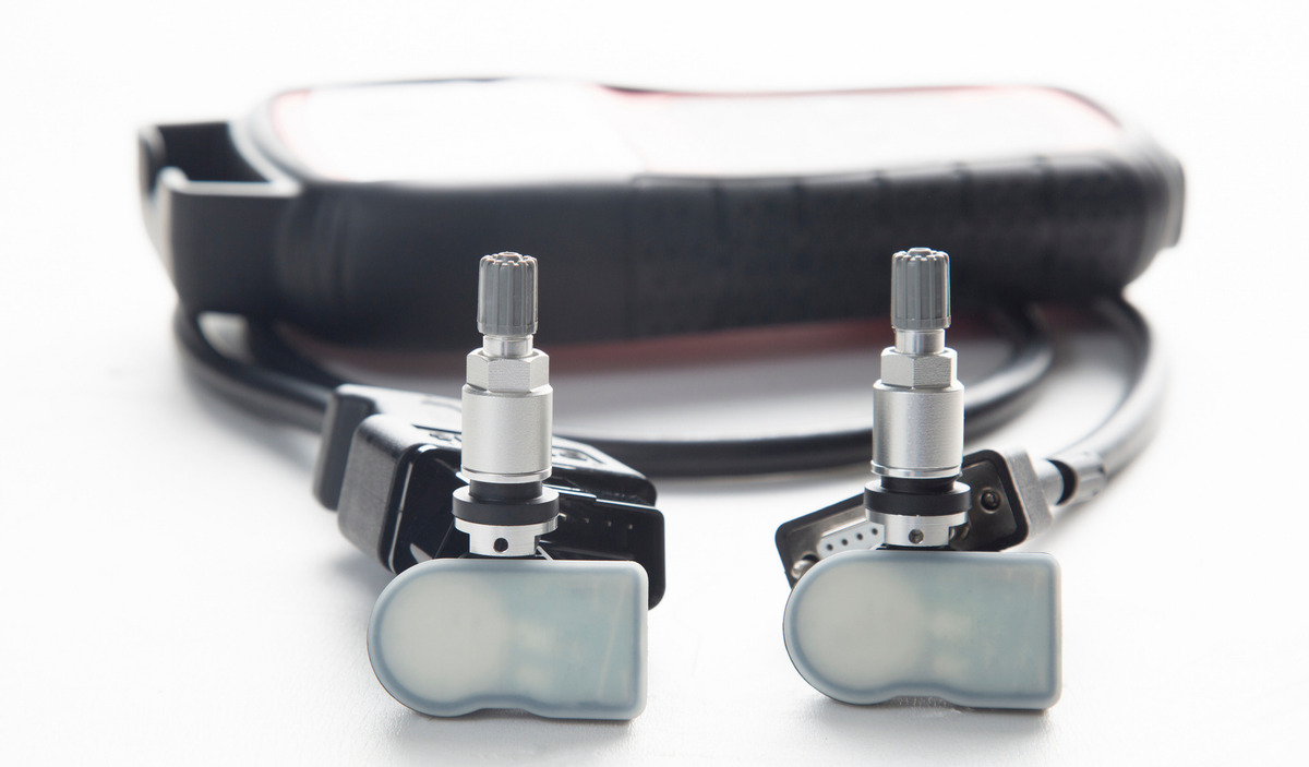 Two white TPMS sensors with aluminum valve stems placed in front of a handheld TPMS programming tool and its cable